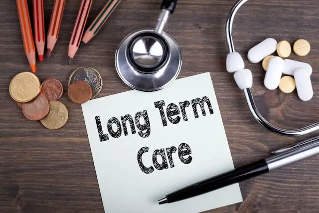 Words 'long term care' written on a piece of paper sitting on a desk next to a pen, stethoscope, money, and pills