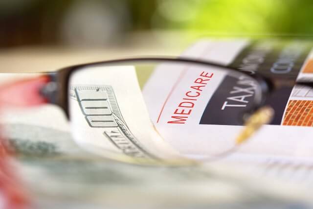 Close up of the word 'medicare' in focus through reading glasses on a desk next to a $100 bill depicting medical/insurance costs