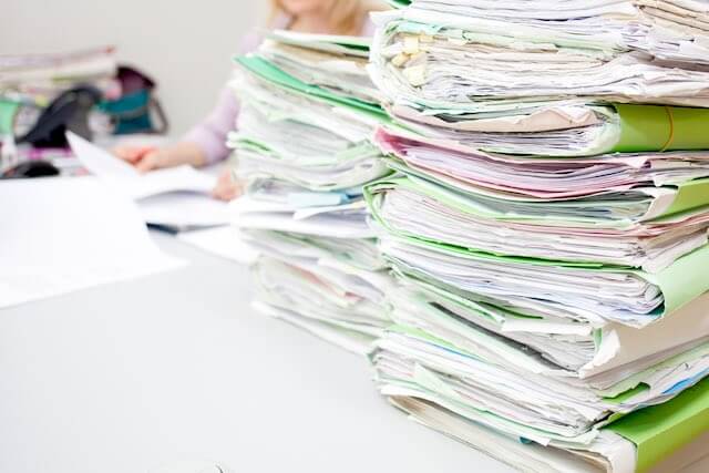 Stacks of file folders filled with papers sitting on a desk with a blurred image of a woman sitting at the desk behind them depicting a paperwork backlog