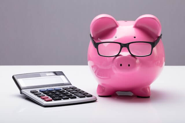 Pink piggy bank wearing glasses next to a calculator on a solid white surface depicting investment planning, returns, fees
