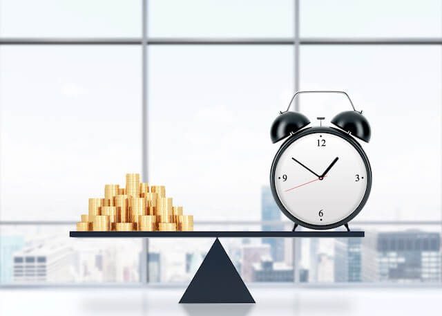 Balance scale with a stack of gold coins on the left and an old style alarm clock on the right depicting official time or a balance of time versus money concept
