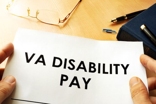 Close up of a person's hands holding a document over a desk that reads 'VA disability pay' with glasses and a pen sitting next to it on the desk