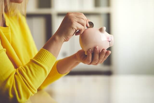 Close up of a woman in a yellow shirt holding a pink piggy bank and putting a coin into it depicting retirement savings