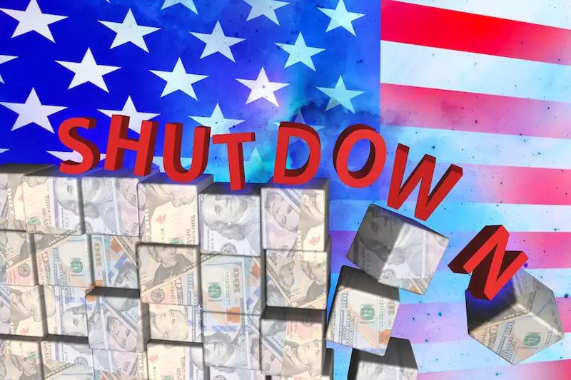 American flag pictured in the background behind blocks of American dollars falling along with the word 'shutdown' depicting a government shutdown and loss of money