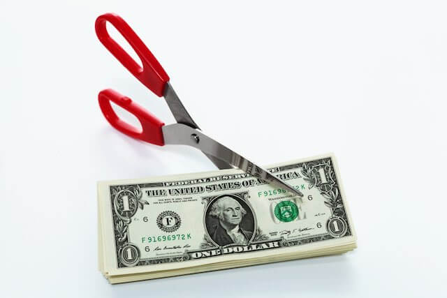 Pair of scissors cutting a stack of dollar bills against a solid white background depicting a pay cut or pay freeze