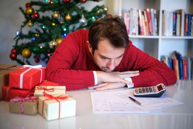 Man sitting at a desk with his chin resting on his hands looking sadly at paperwork on the desk with Christmas gifts next to him and a Christmas tree in the background
