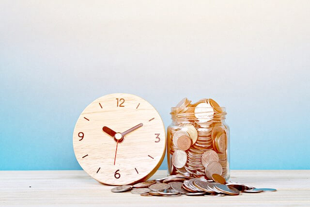 Wooden alarm clock next to a jar of coins depicting time is money concept