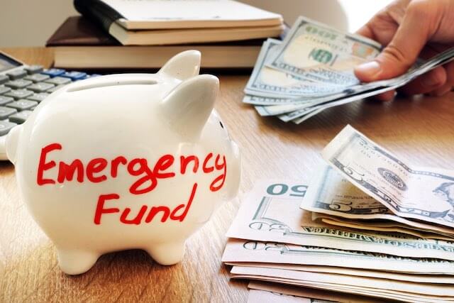 Piggy bank on a desk labeled 'emergency fund' next to stacks of cash and a calculator