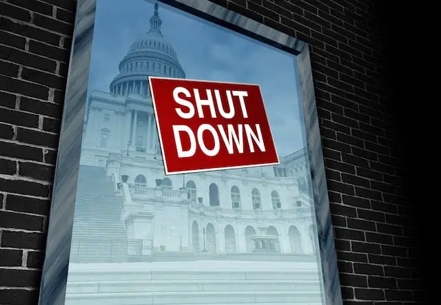Red sign with white letters hanging in a window that reads 'shut down' with a picture of the Washington, DC capitol building visible on the other side of the window