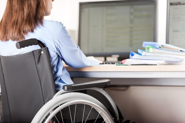 Woman sitting in a wheelchair as she works at her desk on a computer with a stack of paperwork beside her computer workstation - disability retirement, handicapped