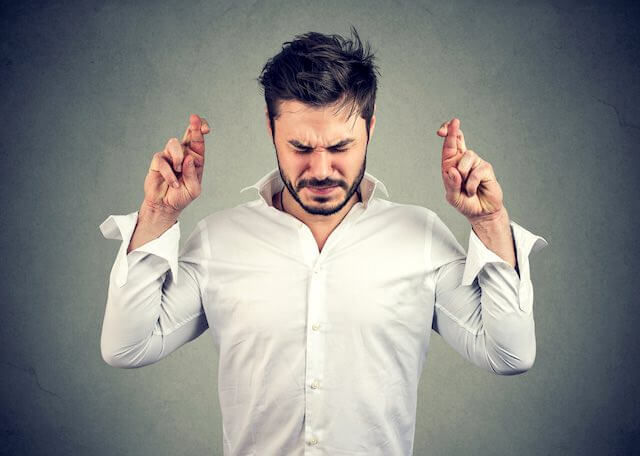 Man standing with his chin bent down, eyes tightly closed, and holding both of his arms up with his fingers crossed appearing to anxiously anticipate or hope for something to happen