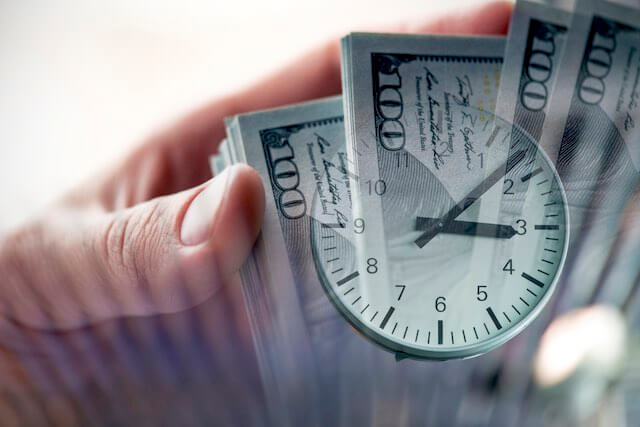 Close up of a person's hand holding a spread of $100 bills with an image of a clock overlaid on top fo the cash depicting back pay, interest payments, time is money