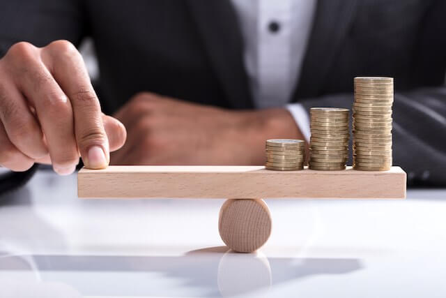 Close up of a businessperson's finger on one side of a scale to balance it against a stack of coins on the other end depicting rebalancing one's investment portfolio