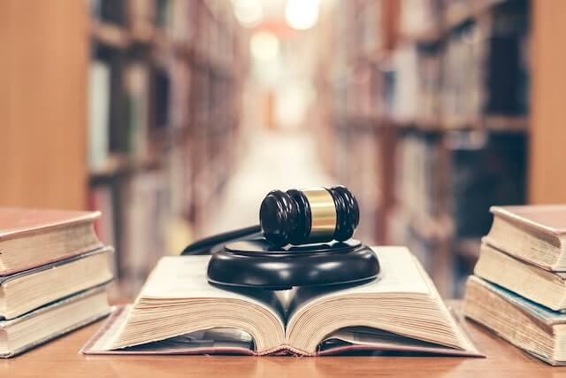 Wooden judge's gavel sitting on top of an open law book on a bookshelf with two stacks of books on either side pictured with a library of bookshelves in the background