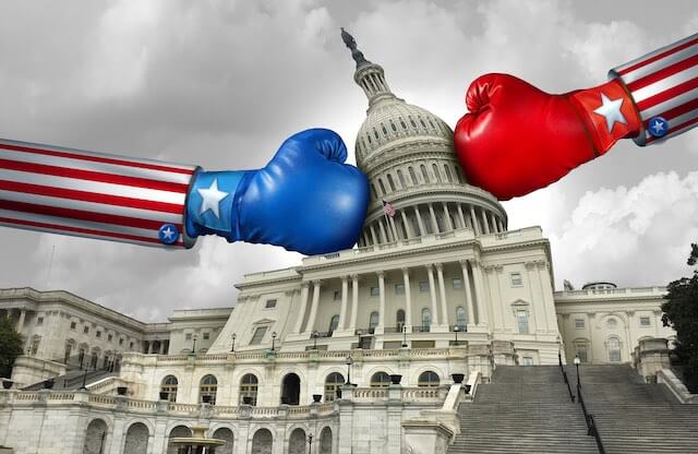 Two boxing gloves coming from both sides, one blue and one red, hitting the Capitol building in Washington, DC depicting a government shutdown fight in Congress