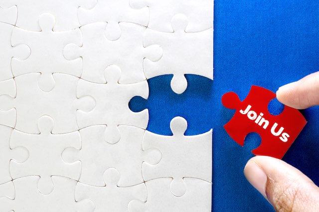 Close up of a person's hand putting a red puzzle piece labeled 'join us' into a missing space in a large puzzle depicting human resources, hiring new employees