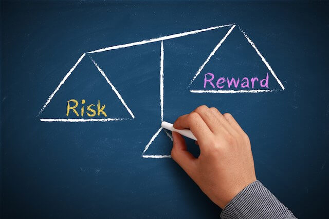 Close up of a person's hand sketching a drawing on a chalkboard of a scale with one side labeled 'risk' and the other side labeled 'reward' depicting risk vs. reward concept in investing, retirement planning