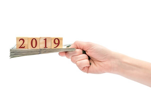 Close up of a woman's hand holding a stack of cash with wooden blocks on top of it that read '2019' depicting a 2019 pay raise