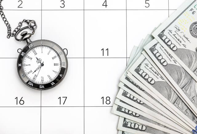 Spread of $100 bills on a calendar next to a pocket watch depicting a payment deadline such as back pay or a future/pending pay raise