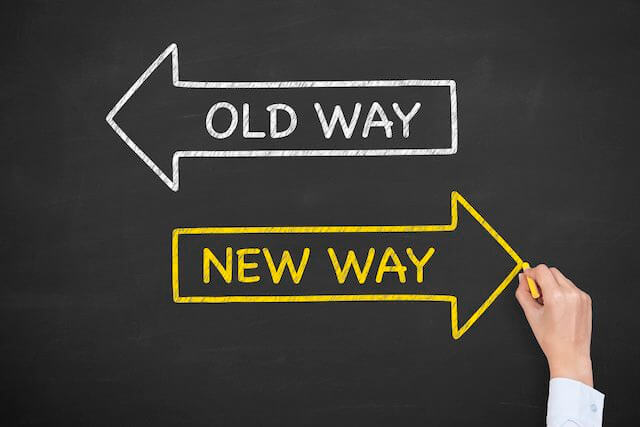 Person's hand drawing two arrows on a chalkboard, one labeled 'old way' in white pointing to the left and one labeled 'new way' in yellow pointing to the right