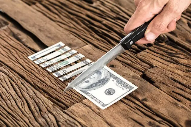 Close up of a person's hand holding a kitchen knife cutting a $100 dollar bill into pieces on a wooden surface depicting pay cuts, budget cuts