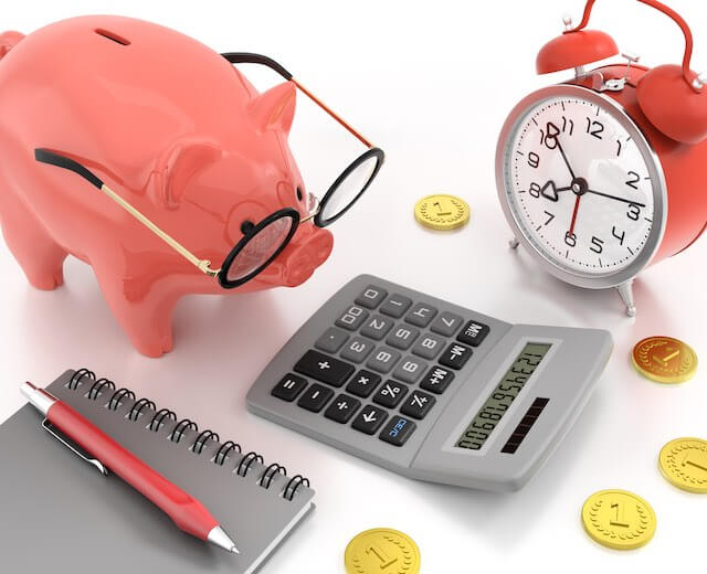 pink piggy bank wearing glasses on a white surface next to a calculator, old style alarm clock, notebook and coins depicting financial planning