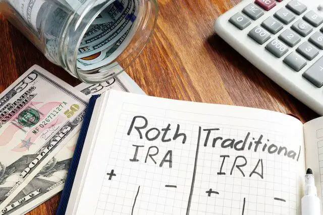 Notebook on a desk with two columns for comparison purposes, one labeled 'Roth IRA', the other labeled 'Traditional IRA' with cash and a calculator next to it