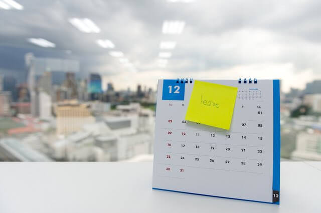 Calendar sitting on a desk with a yellow sticky note pasted on it that has the word 'leave' written on it