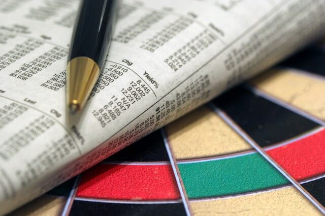 Pen and folded newspaper open to the financial tables section sitting on top of a dartboard depicting investment target allocations