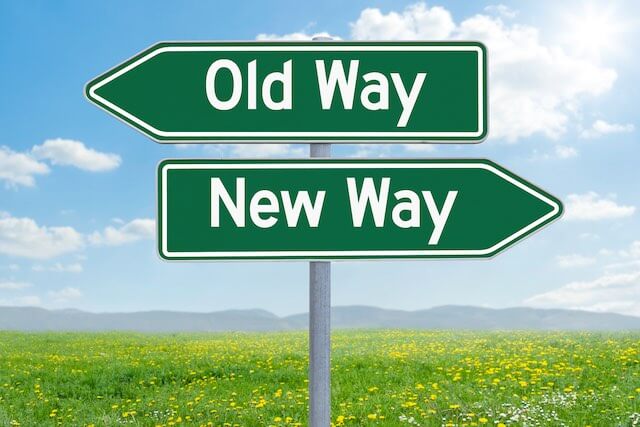 Two green directional road signs with white writing on top of one another pointing in different directions, one reading 'old way' and the other reading 'new way'