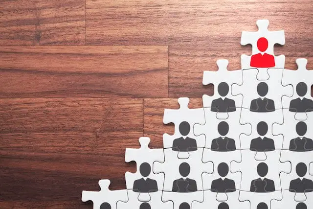 Puzzle pieces on a solid wood surface, each of which has a black colored person icon on it representing employees in an organization hierarchy; the puzzle piece at the top has a red person icon on it depicting the organization's leader
