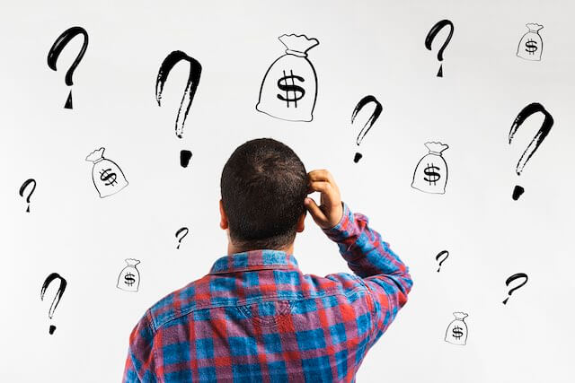 Man with his back facing the viewer scratching his head while looking puzzled as he stares at a whiteboard covered with question mark and money bag icons depicting confusion or questions about money/salaries