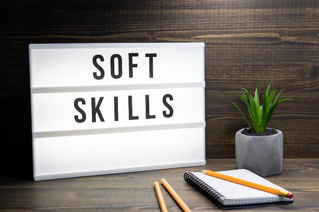 Words 'soft skills' written on a whiteboard sitting on the back of an office desk with pencils and notepad in front of it