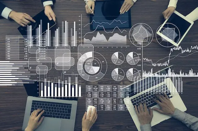 Close up of a group of employees' hands working on laptops sitting around a conference room table with images of graphs and charts overlaid on top of the image depicting business metrics