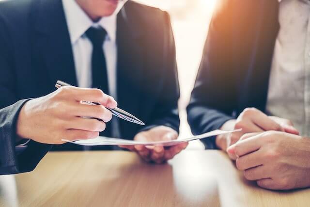 Close up of two businessmen, one of which is holding a pen and pointing it at paperwork he is holding, as they engage in a contract negotiation or business deal