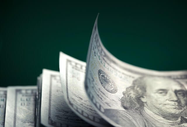 Stack of $100 dollar bills against a solid black background, the top one of which is curled up slightly