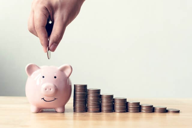 Stacks of coins growing taller as they go from right to left with a pink piggy bank at the end with a closeup of a person's hand putting a coin into it depicting growing wealth and savings for retirement