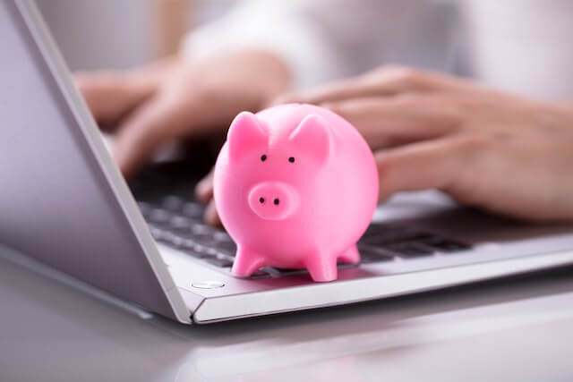 Pink piggy bank sitting on the top of a laptop keyboard next to a closeup of a man's hands typing on the keyboard