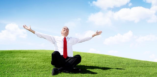 Senior businessman sitting on a grassy hill on a beautiful day with his arms outstretched as he looks toward the sky smiling in a sense of relief and happiness