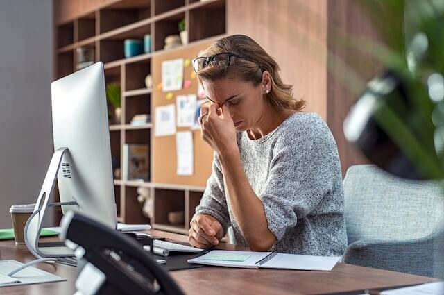 Businesswoman sitting at her desk with her eyes close and her hand pressed against the bridge of her nose as she appears to suffer from a headache and/or stress