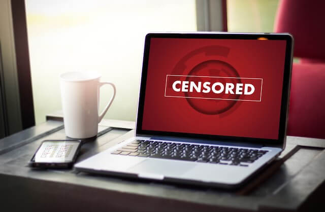 Word 'censored' displayed on a laptop computer screen sitting on a desk next to a smartphone and coffee cup