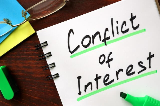 Words 'conflict of interest' written on a notepad on a desk net to reading glasses and a marker