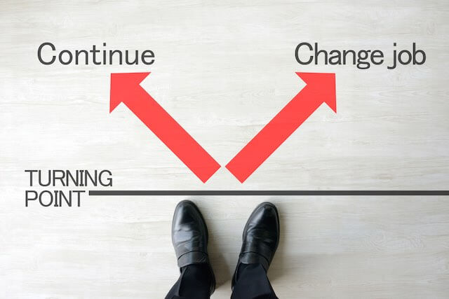 Looking down at a businessman's shoes up against a black line labeled 'turning point' above which lies two red arrows pointing in opposite directions, one labeled 'continue' the other labeled 'change job'