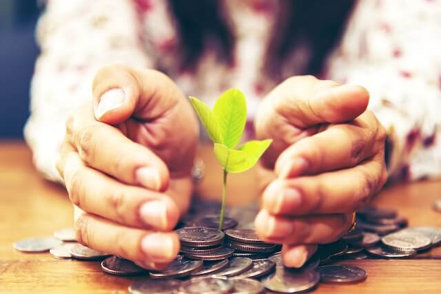 Close up of an elderly woman's hands cupping around a small green plant growing out of a scatting of coins on a wooden desk depicting savings/investing growth for retirement