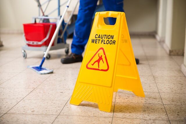 Yellow stand up floor warning sign that says 'caution wet floor' with the feet of a janitorial employee pictured in the background mopping the floor inside of an office building