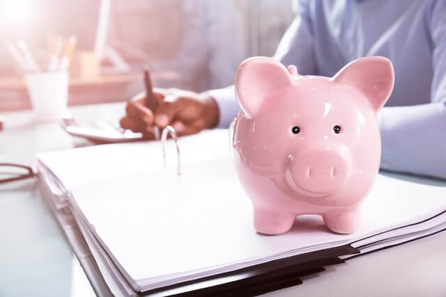 Pink piggy bank sitting on an open workbook binder next to a man at a desk performing financial calculations