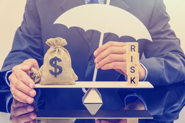 seesaw balance scale with a bag of money on one side and the word 'risk' on the other side with a closeup of a businessman's hands steadying the balance and holding an umbrella over it depicting protecting investments from financial risk