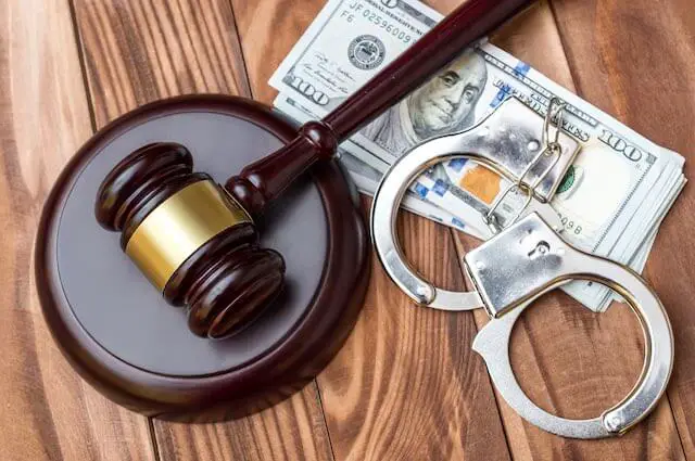 Wooden judge's gavel with a pair of handcuffs and stack of $100 bills on a wooden surface