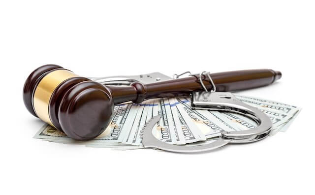 Wooden judge's gavel with handcuffs on top of a spread of cash depicting a crime or guilty plea in court