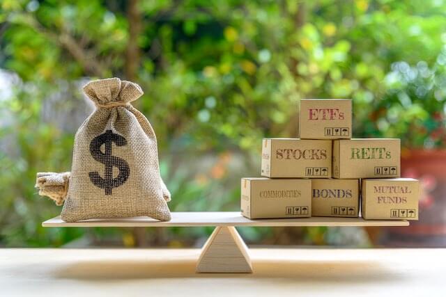 See-saw balance scale with a bag of money on one side and a stack of blocks on the other; individual blocks are labeled 'ETFs', 'stocks', 'REITs', 'commodities', 'bonds', and 'mutual funds' depicting investment diversification